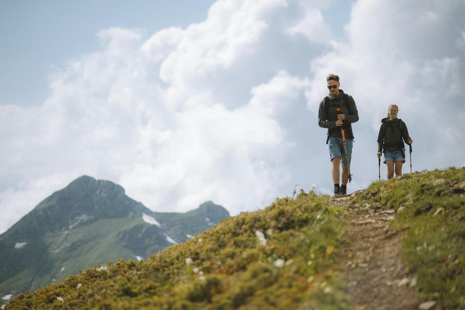 A couple enjoying a hike through lush mountains with trekking poles under a cloudy sky.